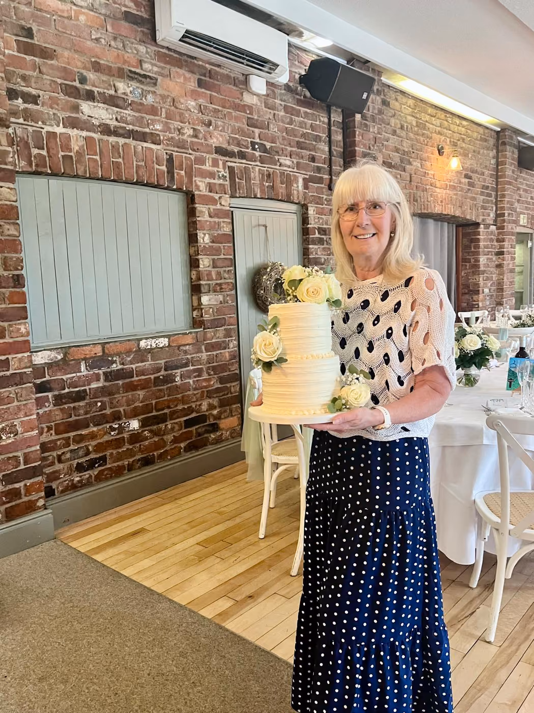 Geraldine holding a two-tier white buttercream wedding cake with white roses at a wedding venue