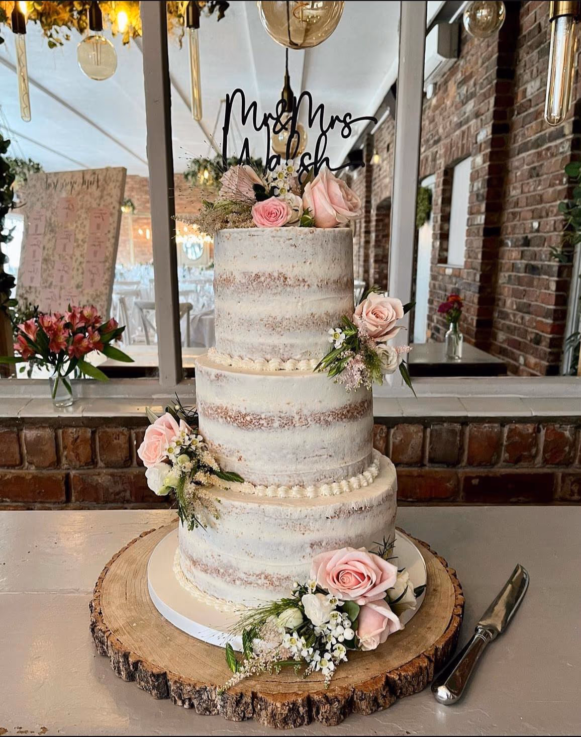 Three-tier naked wedding cake with pink roses, greenery, and wood slice