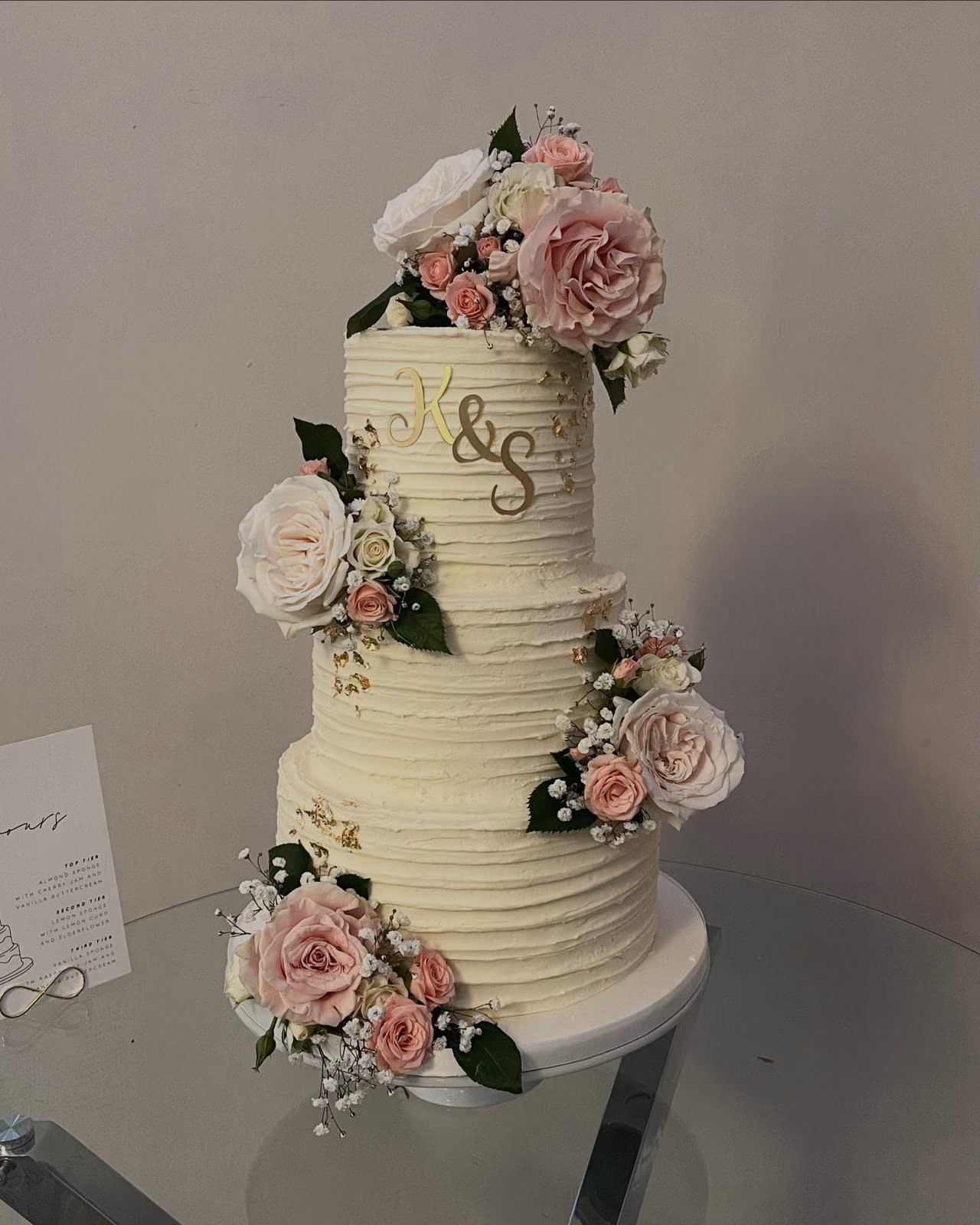 Three-tier textured buttercream wedding cake with pink and white roses, gold leaf, and a gold monogram